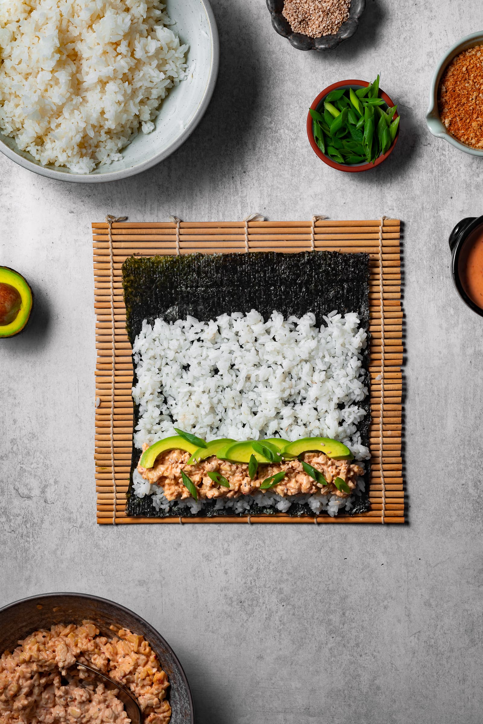 Sushi rice on a sheet of nori with a thin strip of tempeh tuna filling, avocado, and green onions.