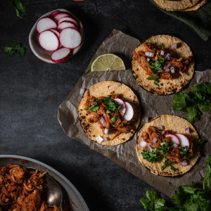 Three tacos with toppings on a parchment paper surrounded by more taco fixings.