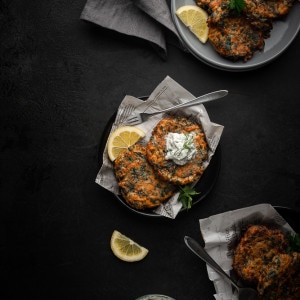 3 plates filled with vegetable fritters with a dollop of vegan tzatziki sauce on top with fresh dill, lemon wedges, and parsley