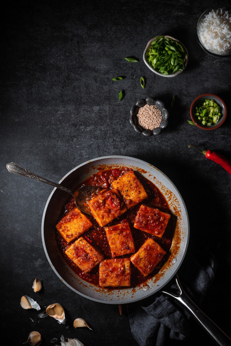 Braised tofu in a frying pan surrounded by serving ingredients like scallions, rice, sesame seeds.