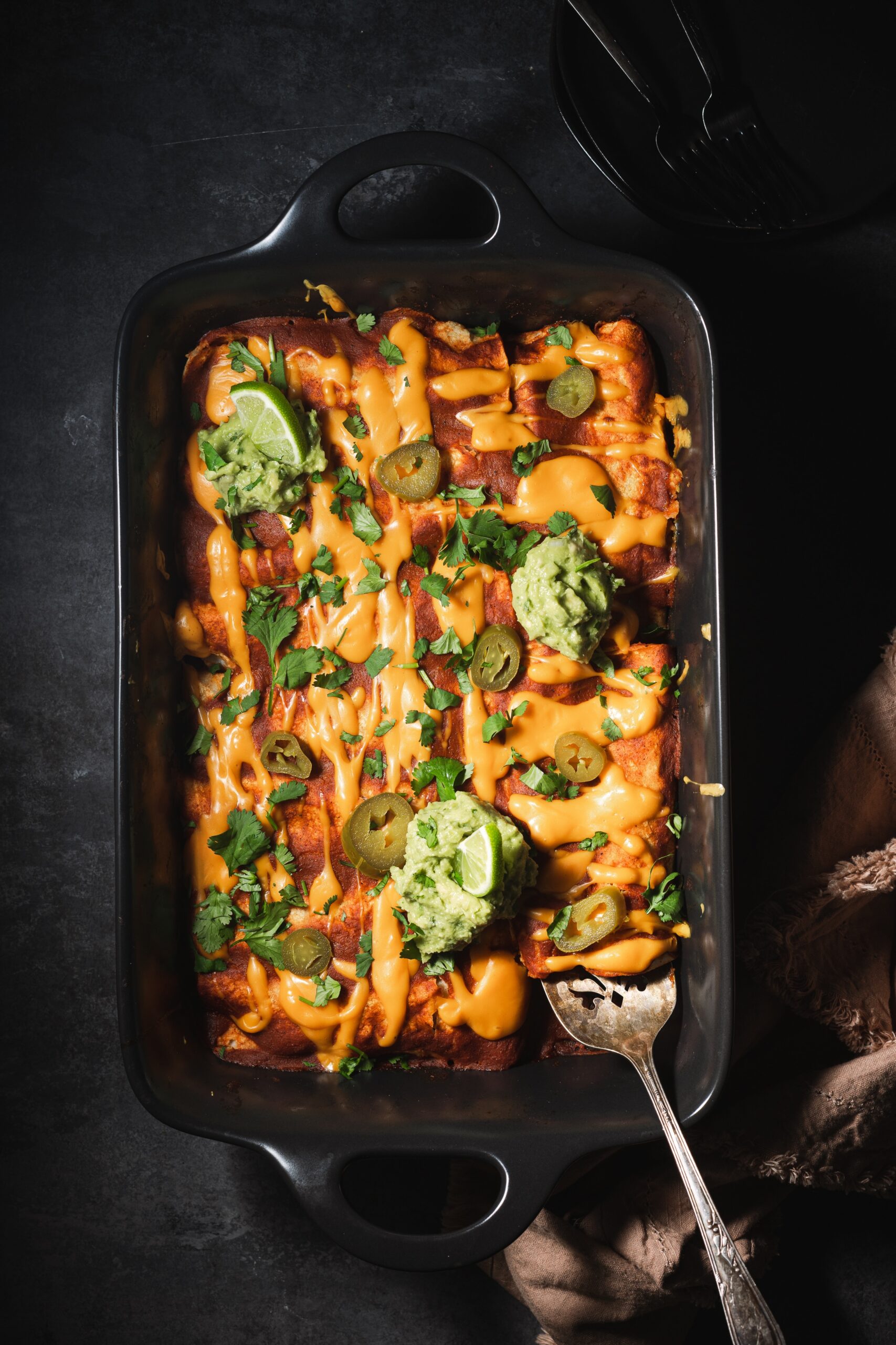 Roasted vegetable and black bean enchiladas in baking tray garnished with guacamole, lime wedges, and jalapeños.