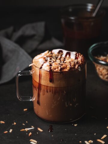 Clear mug filled with a Mocha Latte topped with chocolate syrup drizzle and toasted coconut.
