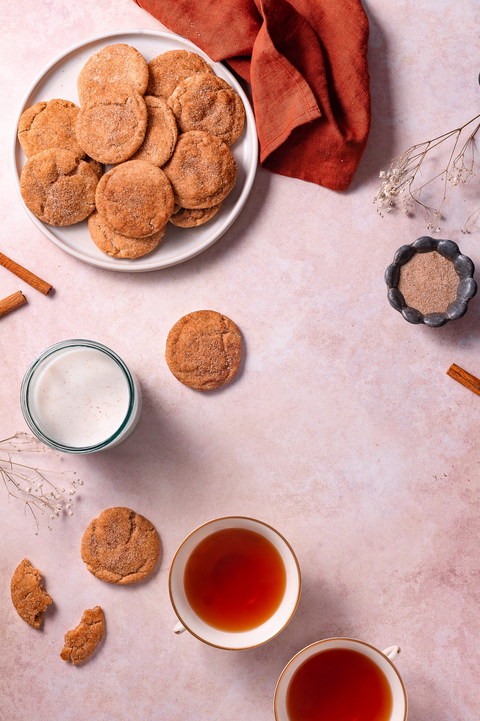 Snickerdoodle cookies on a plate surrounded by cinnamon sticks, glass of milk, and cup of tea.