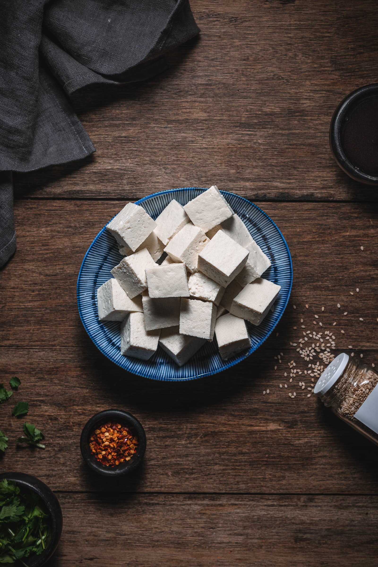 Raw tofu placed in a blue dish surrounded by chili flakes, cilantro and sesame seeds.