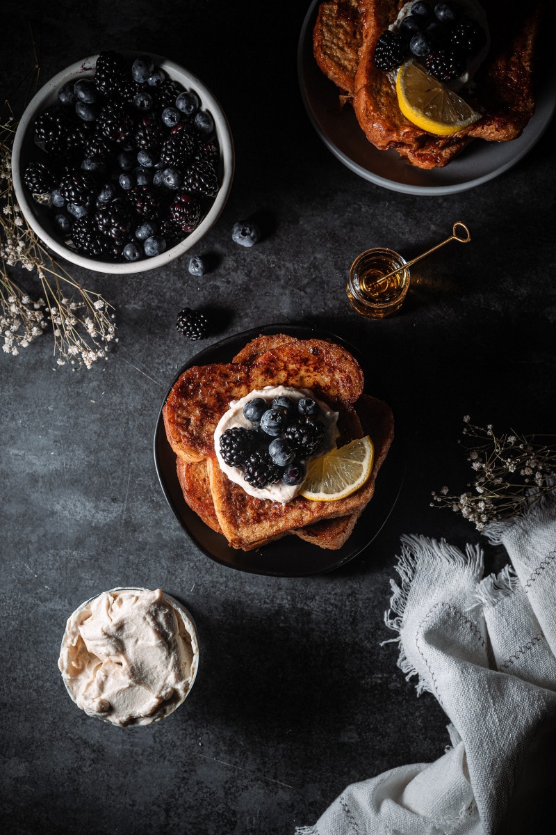 Four pieces of french toast stacked on a plate and topped with vegan mascarpone and blueberries and blackberries.