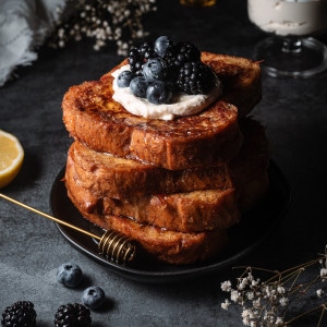 Four pieces of french toast stacked on a plate and topped with vegan mascarpone and blueberries and blackberries.