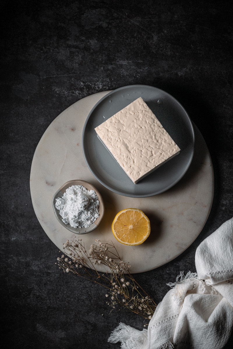 Tofu, powdered sugar and a lemon slice sitting on a marble cutting board.