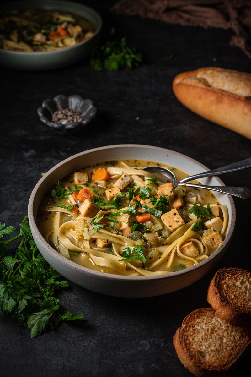 A serving of vegan chicken noodle soup in a serving bowl with bread.