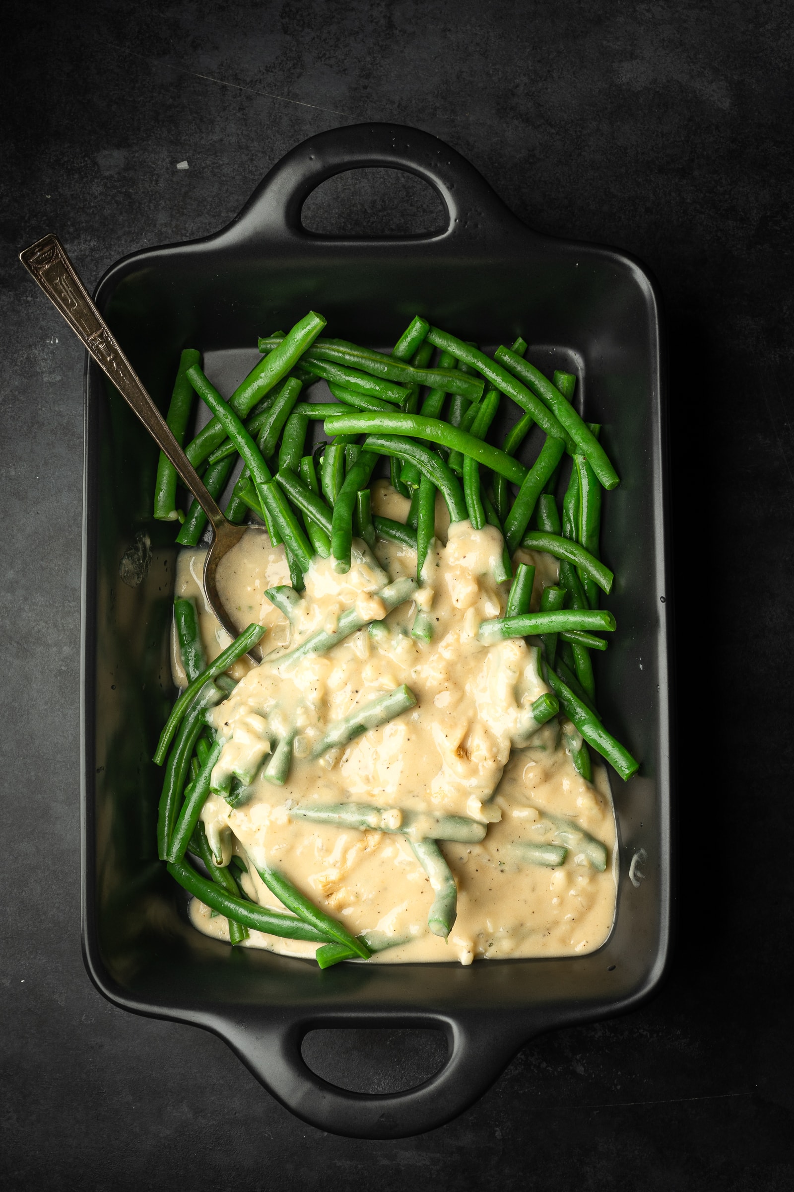 Green beans being mixed with creamy mixture in a black casserole dish.
