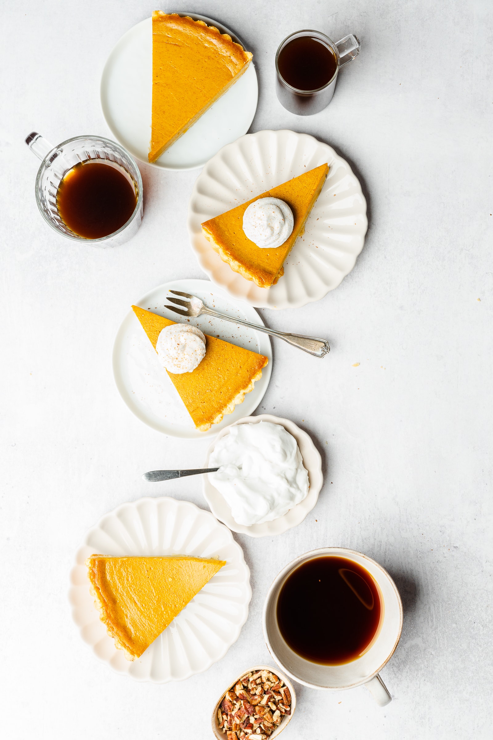 Multiple servings of pumpkin pie on small plates with coffee and side of whipped cream and pecans.