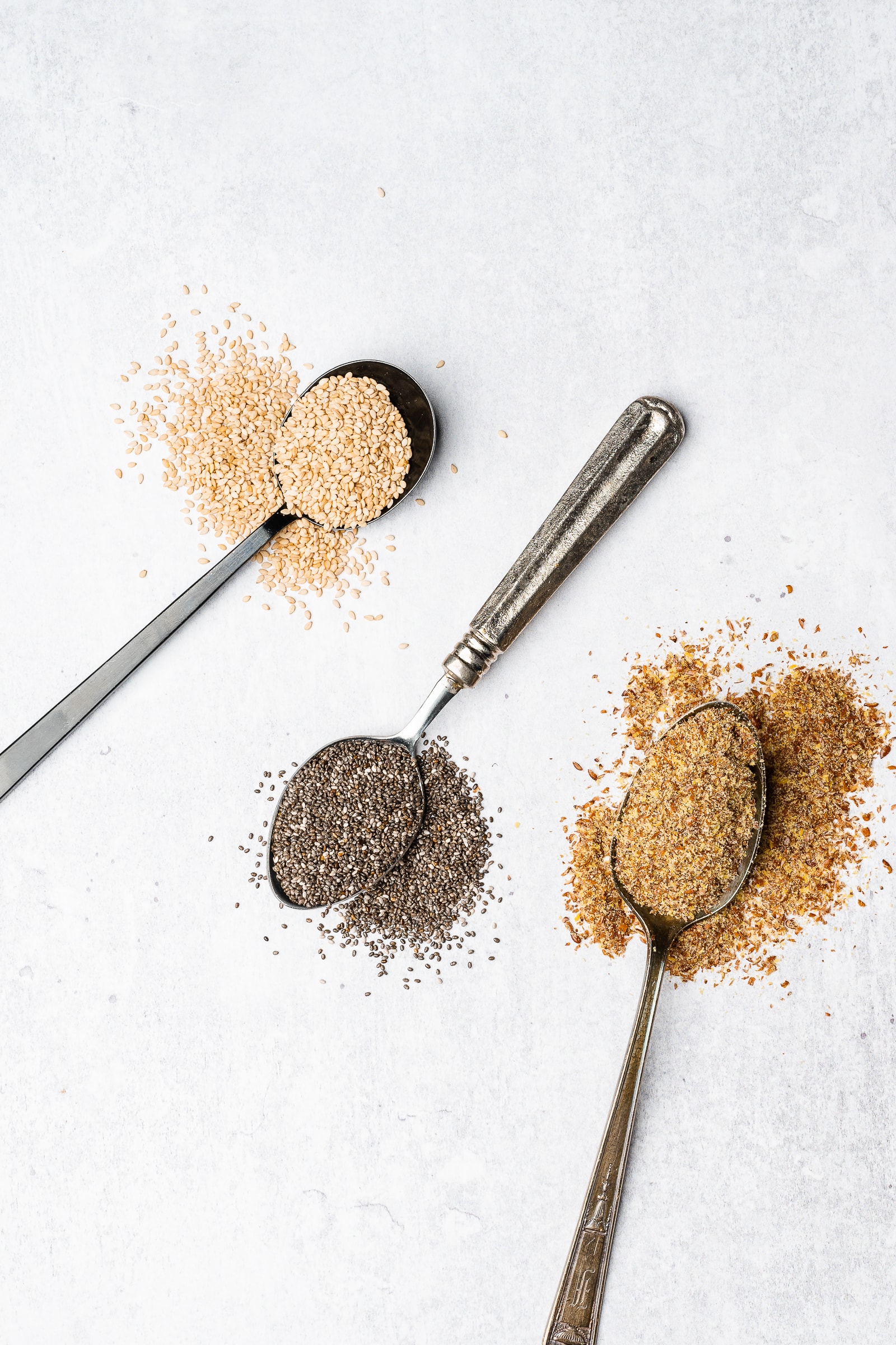 Flaxseed, chia seeds, and sesame seeds on spoons spilled out on a gray/white backdrop.