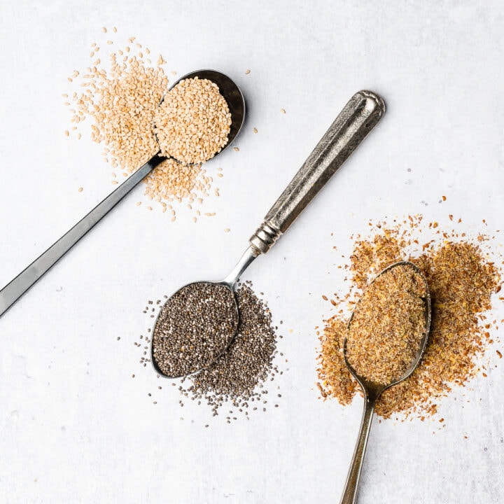 Flaxseed, chia seeds, and sesame seeds on spoons spilled out on a gray/white backdrop.