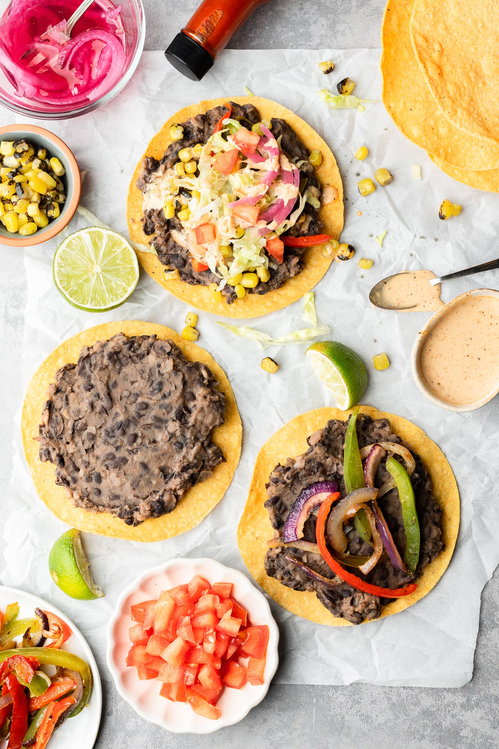 Vegan tostadas topped with black beans and fajita veggies on a piece of parchment surrounded by toppings.
