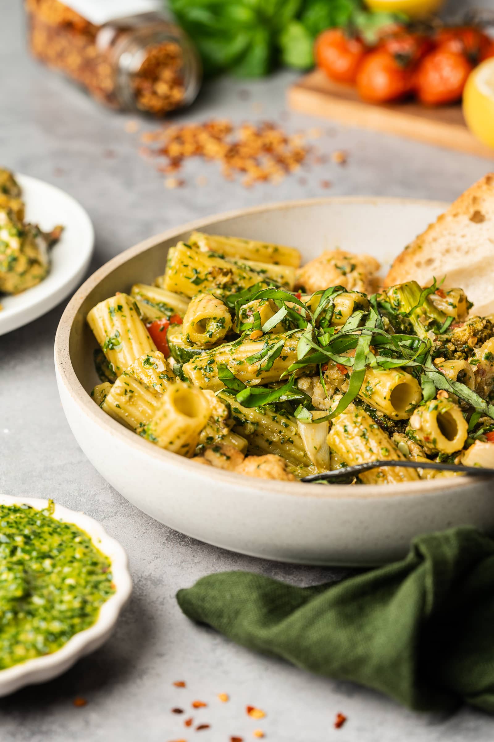 Pesto pasta in a serving dish with bread, roasted tomatoes, fresh basil, and red chili flakes.