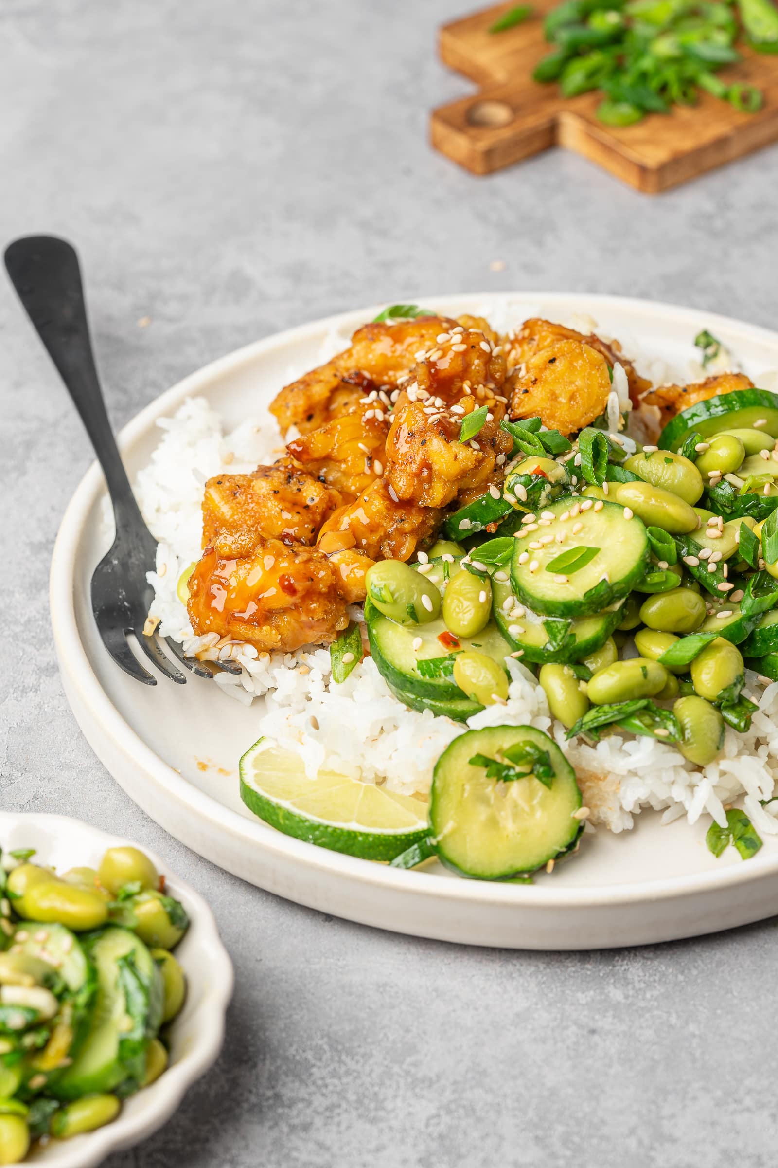 Teriyaki tempeh plated over rice with edamame cucumber salad and garnished with sesame seeds.