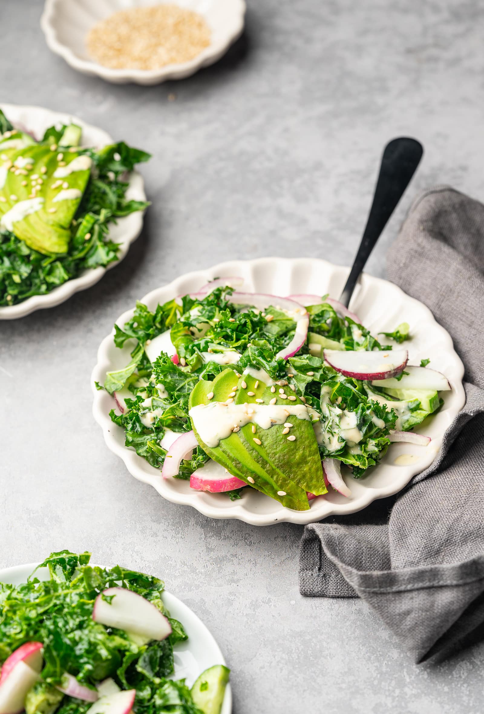Kale salad with tahini dressing served on plates with avocado slices and sesame seeds.