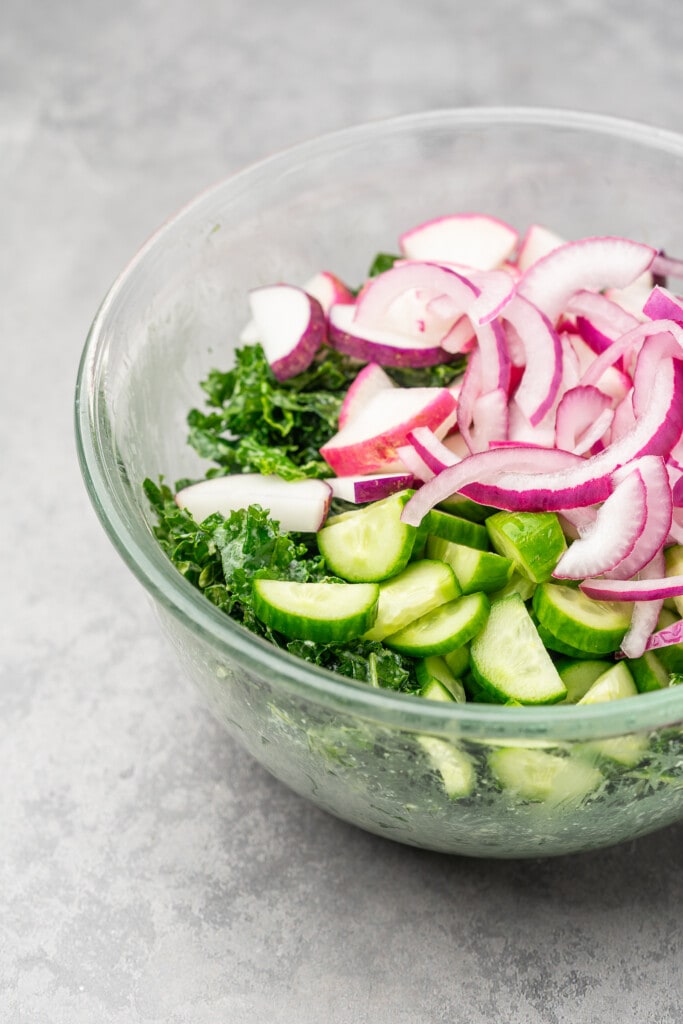 Massaged kale in glass bowl with red onion, radish, and cucumber.