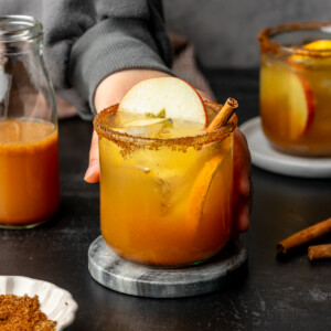 Apple cider mocktail in a glass cup garnished with apple, orange, and cinnamon stick on a marble coaster.