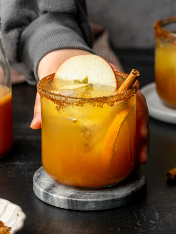 Apple cider mocktail in a glass cup garnished with apple, orange, and cinnamon stick on a marble coaster.