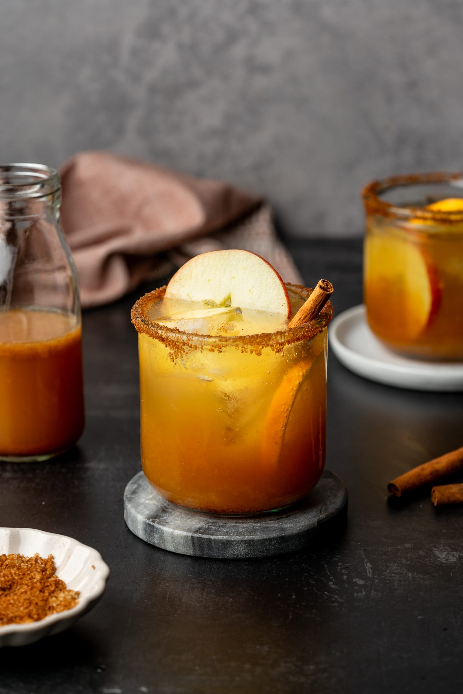 Apple cider mocktail in a glass cup garnished with apple, orange, and cinnamon stick on a marble coaster.