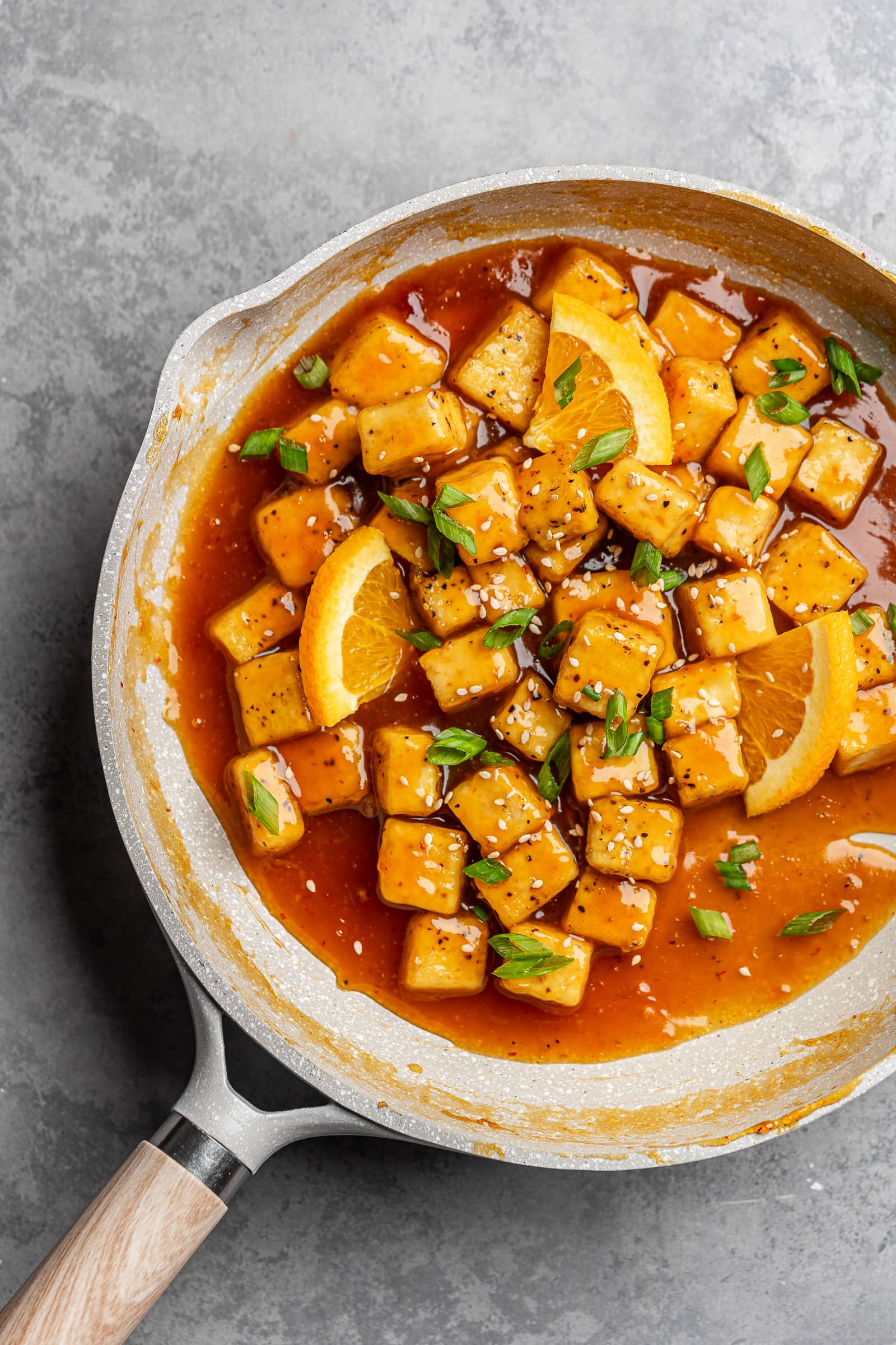 Orange tofu in a pan and garnished with sesame seeds and green onions.