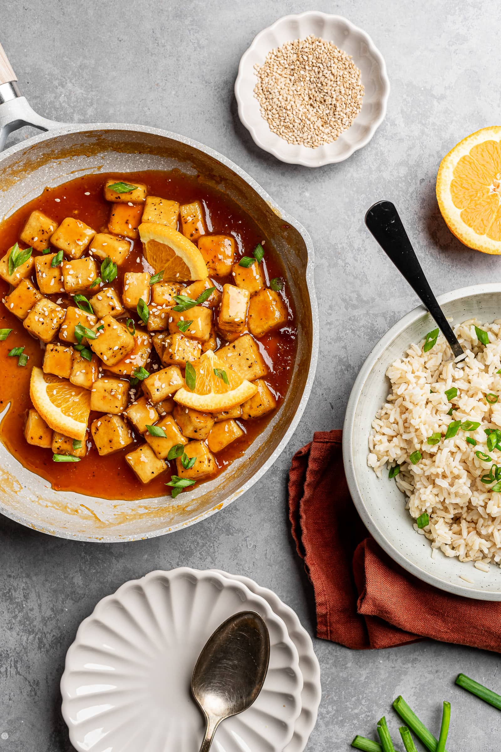 Orange tofu in a pan with a side of rice and garnished with sesame seeds and green onions.