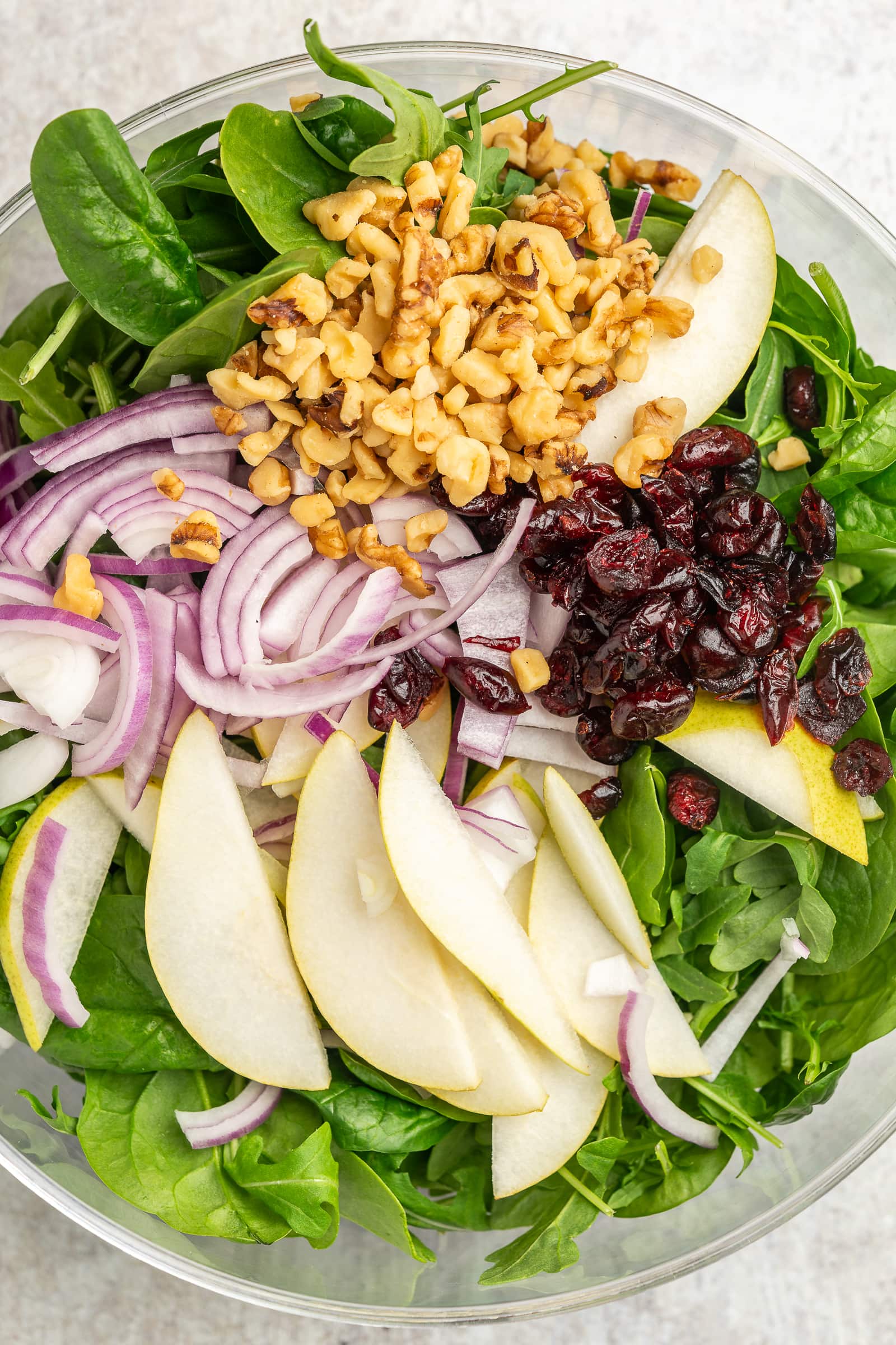 Arugula and baby spinach mixed together in a large bowl topped with pear slices, dried cranberries, walnuts, and red onion.