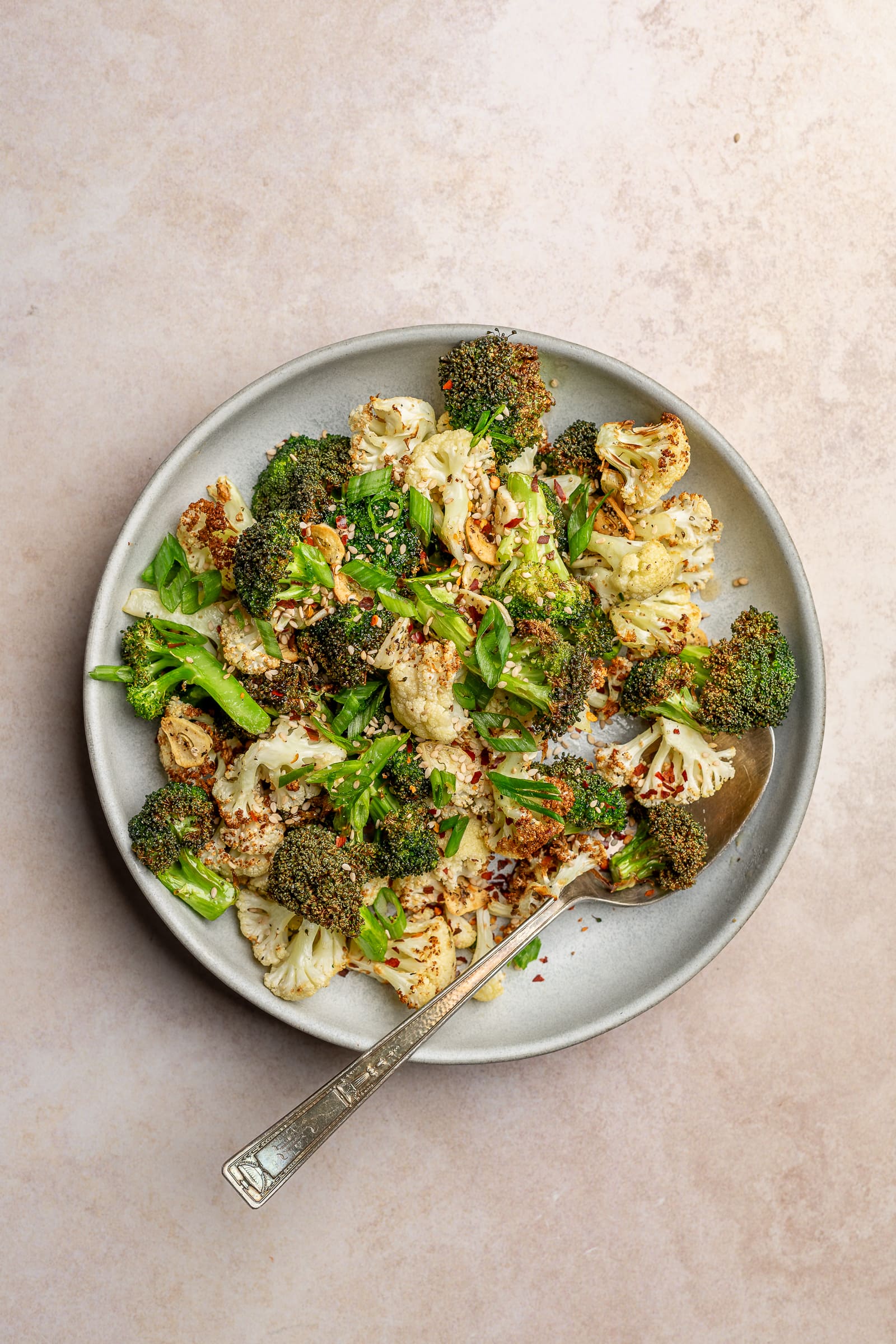 Close up of crispy broccoli and cauliflower with sesame seeds, chili flakes, and green onions on a serving plate.