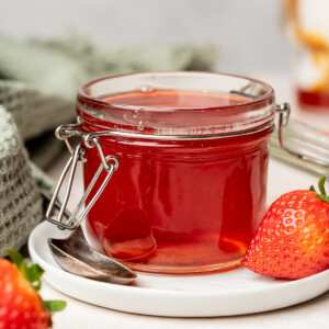 Strawberry simple syrup in a small jar on a white plate and surrounded by strawberries.