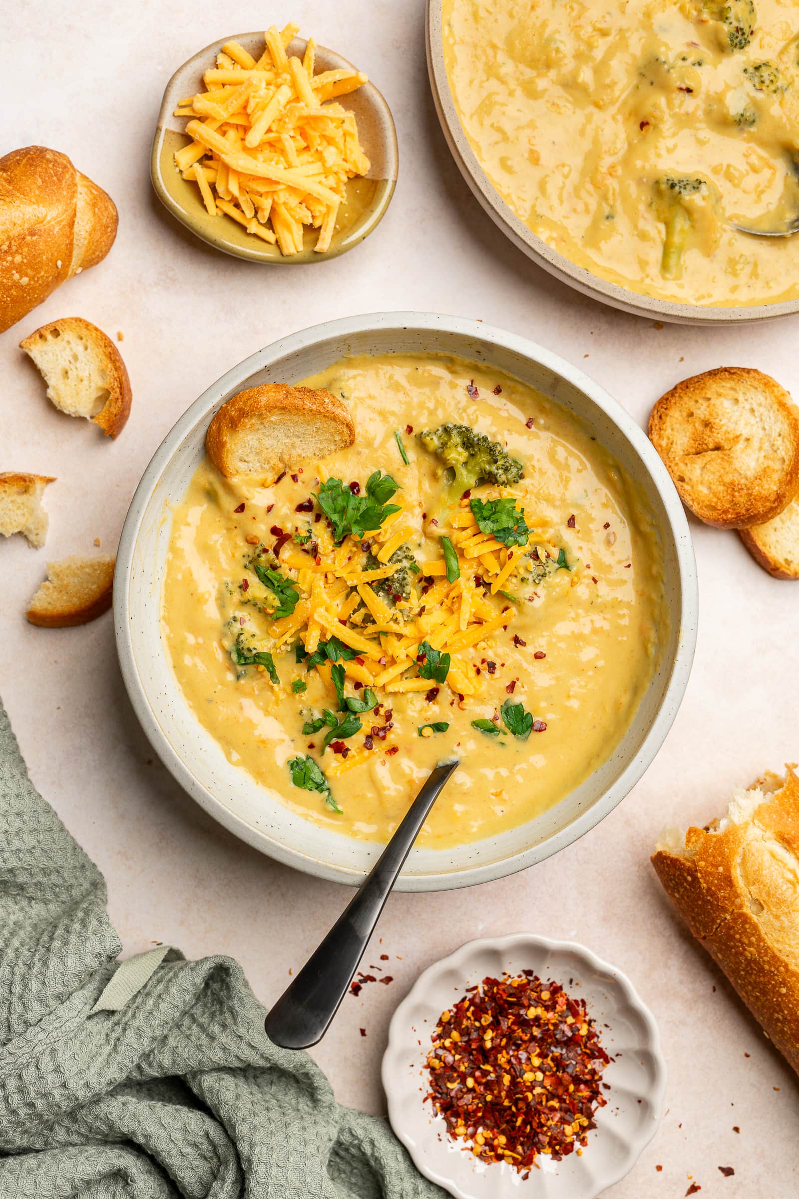 Broccoli cheese soup garnished with cheese, parsley, and red chili flakes with bread on the side.