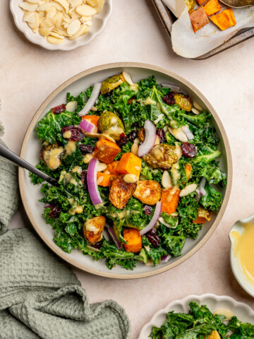 Kale salad in a serving bowl topped with roasted sweet potatoes, brussels sprouts, red onion, and dried cranberries.