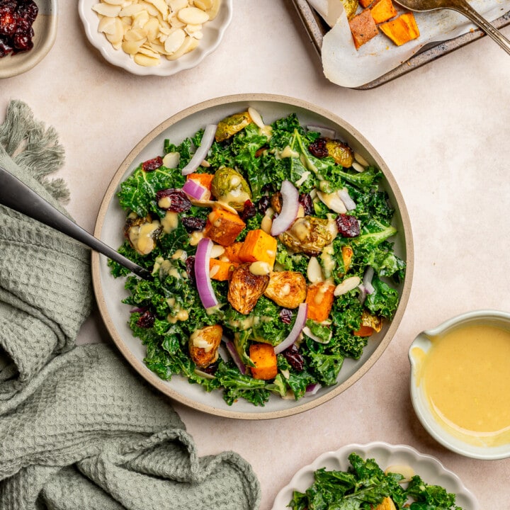 Kale salad in a serving bowl topped with roasted sweet potatoes, brussels sprouts, red onion, and dried cranberries.