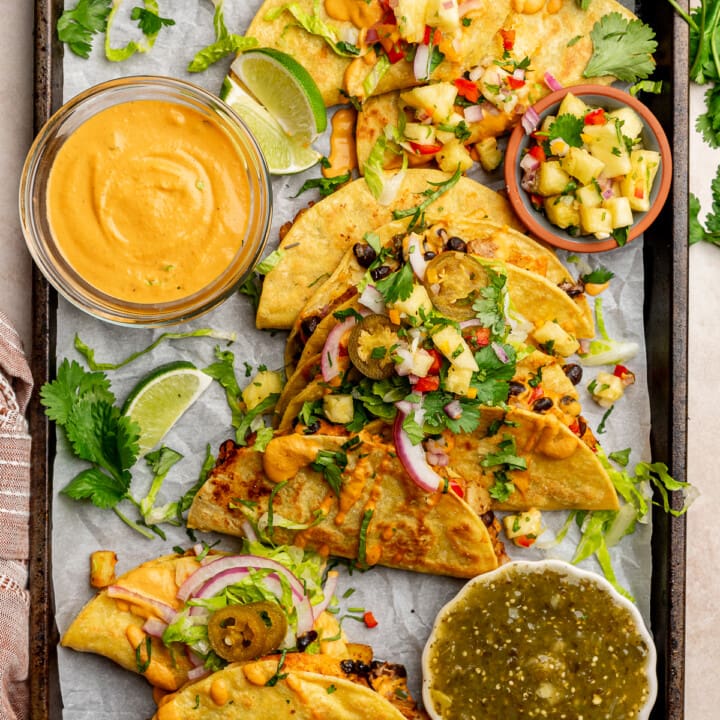 Crispy black bean tacos on a baking tray with various salsas and toppings.