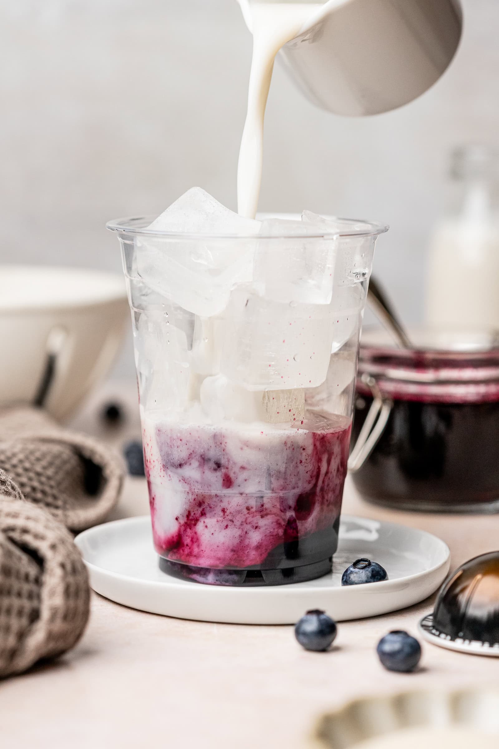 Ice and blueberry syrup in a plastic coffee cup with oat milk being poured in.
