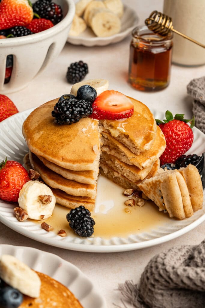 Stack of oat milk pancakes with fresh fruit on top and a drizzle of maple syrup with a bite cut out.