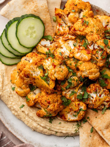 Harissa cauliflower plated on top of hummus and garnished with herbs, served with cucumber and pita bread.