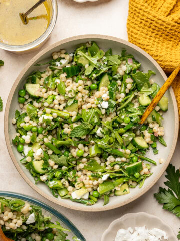 Vegan couscous salad with spring vegetables, vegan feta, and a lemon vinaigrette served in a large bowl with a garnish of fresh mint and parsley.