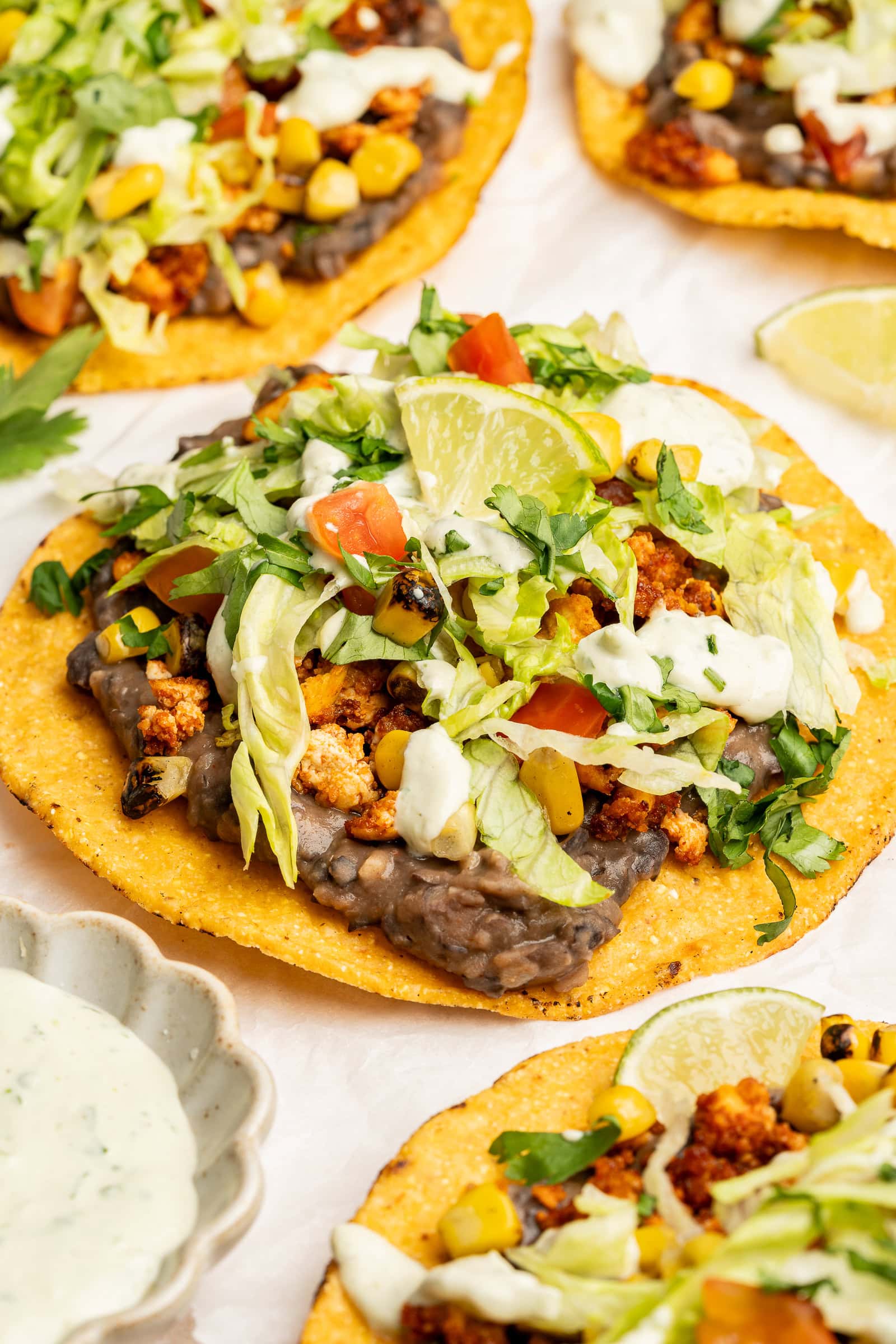 Close up of tofu tostada shell with lettuce, tomato, corn, cilantro crema, and lime wedge.