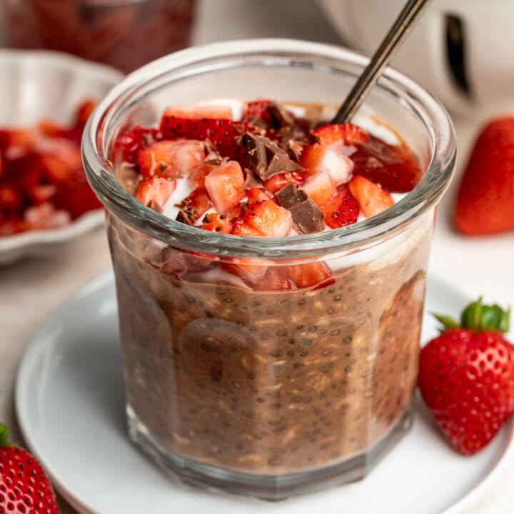 A bowl of chocolate overnight oats with strawberries and a spoon garnished with strawberry jam and chocolate shavings.