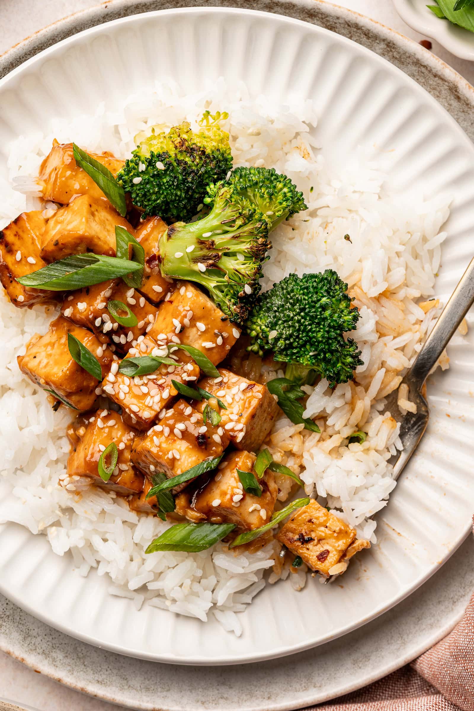 Miso glazed tofu on top of a bed of rice with charred broccoli, sesame seeds, and green onions being eaten with a fork.