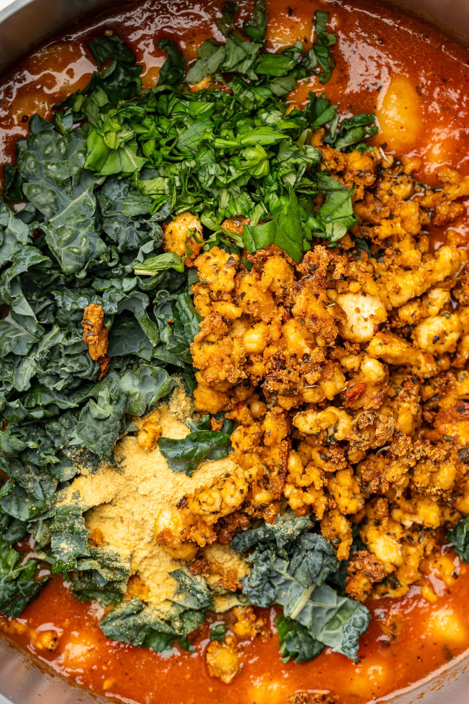 Seasoned crispy tofu, nutritional yeast, chopped kale, and chopped basil being added to gnocchi tomato soup.