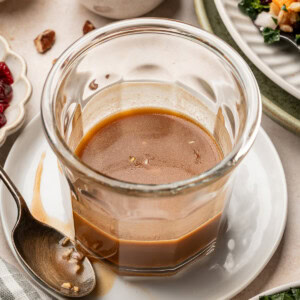 A glass jar of balsamic maple dressing sitting on a plate, surrounded by a kale salad and chopped pecans.