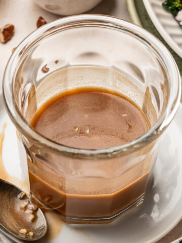A glass jar of balsamic maple dressing sitting on a plate, surrounded by a kale salad and chopped pecans.