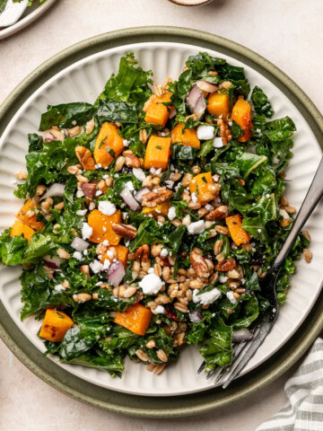 A serving plate of butternut squash kale salad topped with crumbled feta, red onion slices, and pecans.