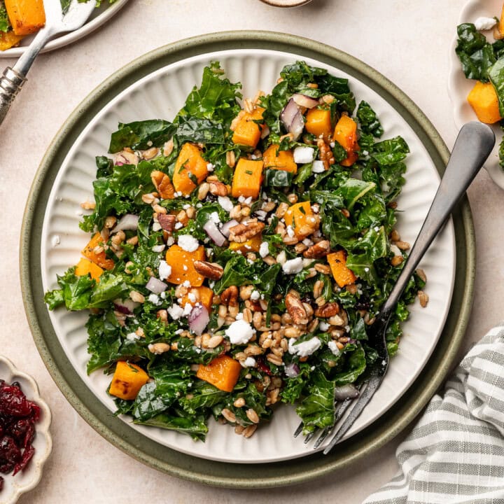 A serving plate of butternut squash kale salad topped with crumbled feta, red onion slices, and pecans.