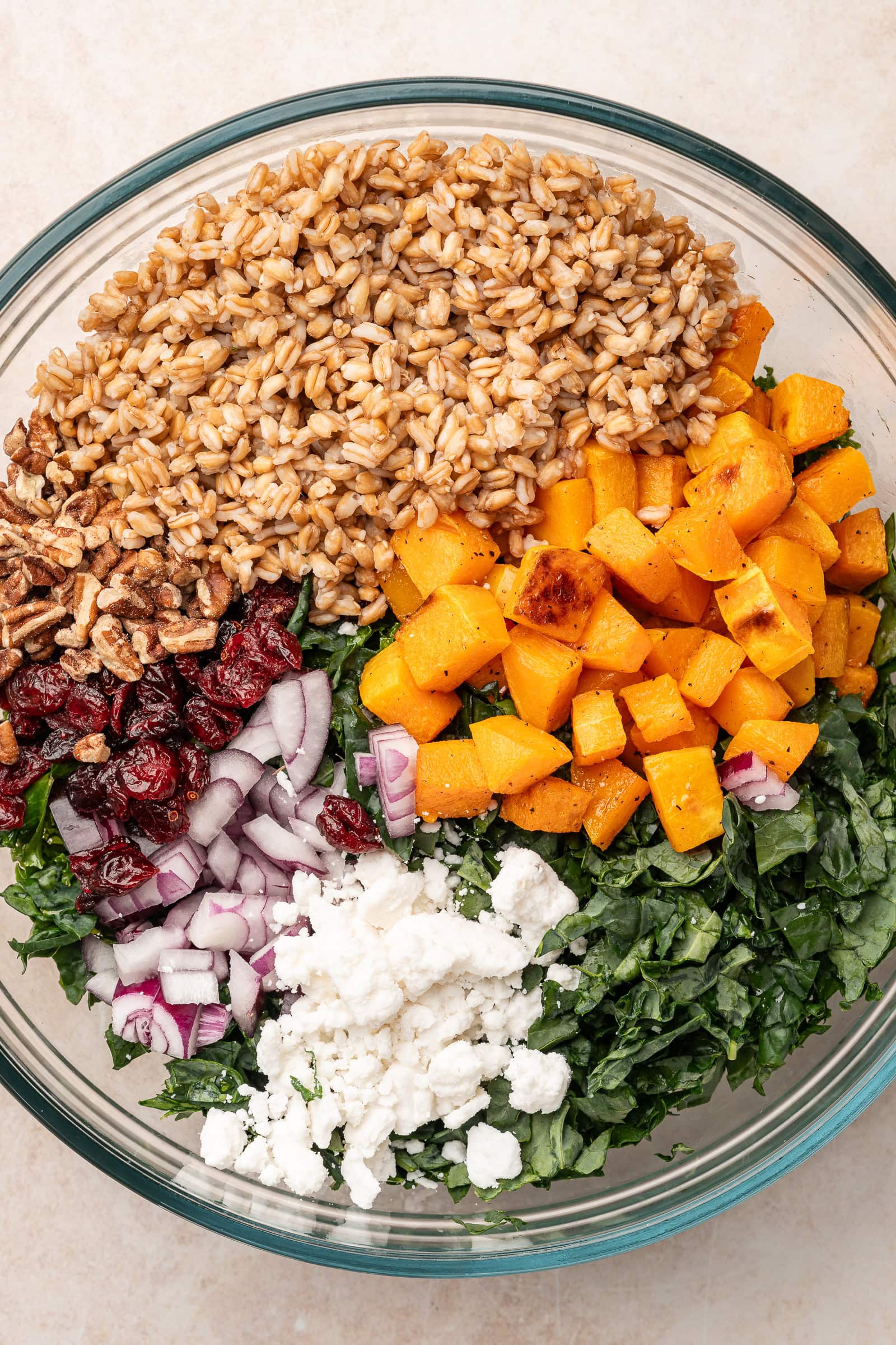 Roasted squash, cooked farro, crumbled feta, diced red onion, dried cranberries, and chopped kale in a large glass bowl.