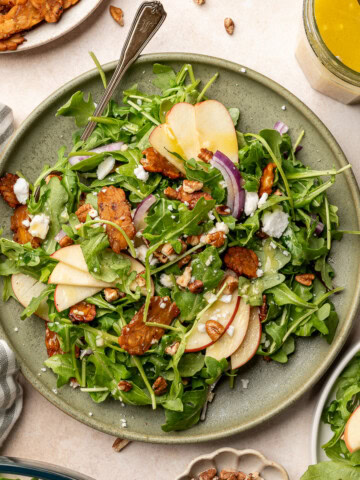 Apple arugula salad garnished with red onion, tempeh bacon, and vegan feta on a serving plate.
