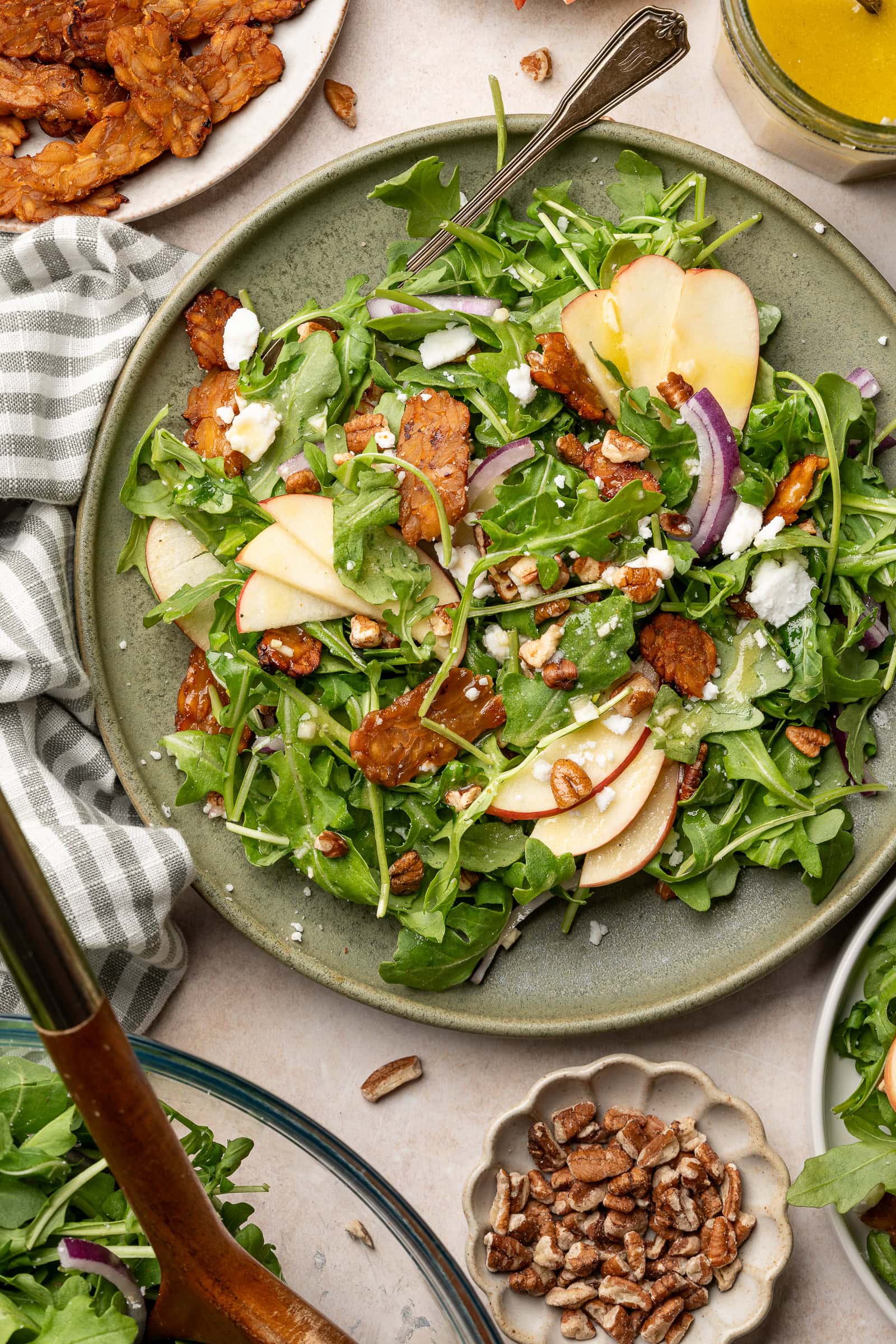 Apple arugula salad garnished with red onion, tempeh bacon, and vegan feta on a serving plate.