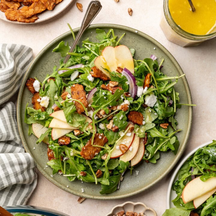 Apple arugula salad garnished with red onion, tempeh bacon, and vegan feta on a serving plate.