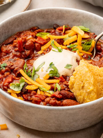 Three bean tvp chili in a serving bowl with a dollop of sour cream, vegan cheddar cheese shreds, and herbs along with a piece of cornbread.